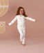 Young child wearing white long-sleeve pyjamas with a colorful all-over print, standing barefoot against a soft pink background with arms outstretched.