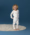 Young child wearing white long-sleeve pyjamas with a playful multicolour print, standing barefoot on a rug against a deep blue background and smiling at the camera.