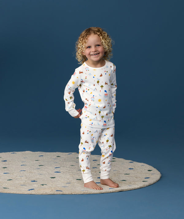 Young child wearing white long-sleeve pyjamas with a playful multicolour print, standing barefoot on a rug against a deep blue background and smiling at the camera.