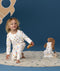 Young child wearing white long-sleeve pyjamas with a colourful illustrated print, kneeling barefoot on a rug against a deep blue background, holding a toy while a teddy bear in matching sleeping bag sits nearby.