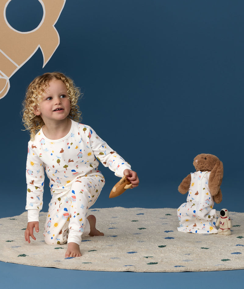 Young child wearing white long-sleeve pyjamas with a colourful illustrated print, kneeling barefoot on a rug against a deep blue background, holding a toy while a teddy bear in matching sleeping bag sits nearby.