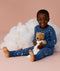 Young child wearing blue Long Sleeve Pyjamas with a moon-and-star print, sitting barefoot on a soft pink background, smiling and holding a teddy bear in front of a fluffy cloud prop.