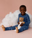 Young child wearing blue Long Sleeve Pyjamas with a moon-and-star print, sitting barefoot on a soft pink background, smiling and holding a teddy bear in front of a fluffy cloud prop.