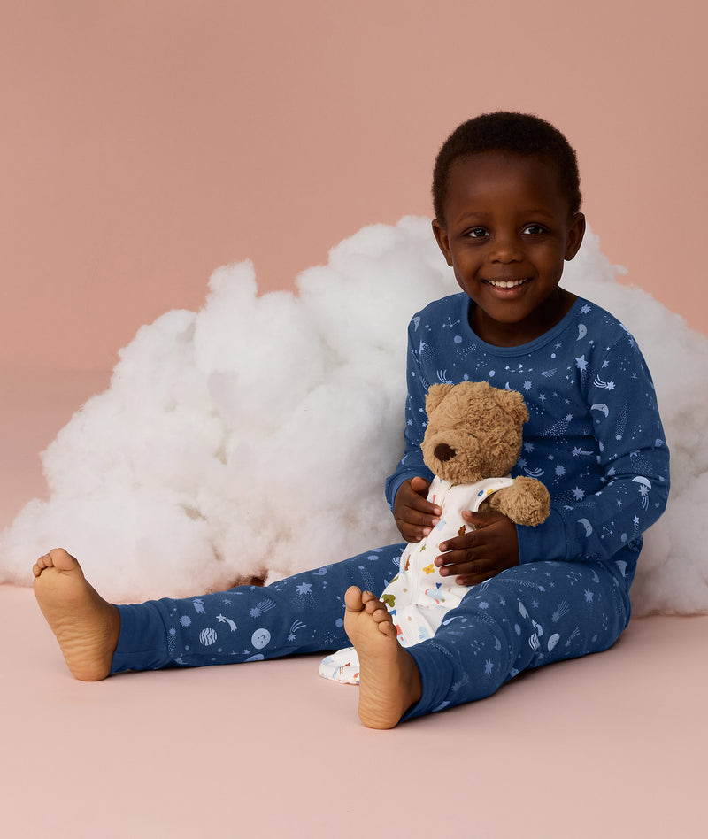 Young child wearing blue Long Sleeve Pyjamas with a moon-and-star print, sitting barefoot on a soft pink background, smiling and holding a teddy bear in front of a fluffy cloud prop.