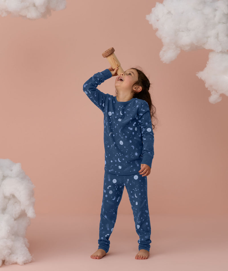 Preschooler wearing blue Long Sleeve Pyjamas with a moon-and-star print, standing barefoot against a soft pink background and looking upward while holding a toy telescope, surrounded by fluffy cloud props.