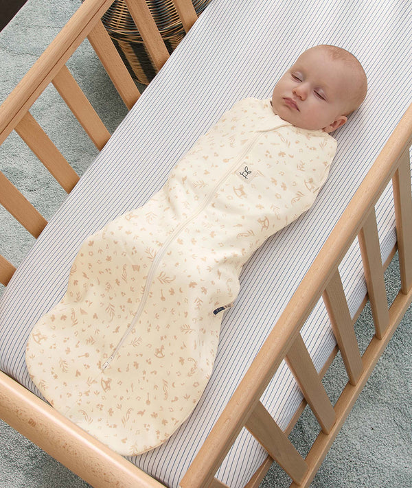Sleeping baby swaddled inside a cream-colored patterned Cocoon Swaddle Bag in a wooden cot.