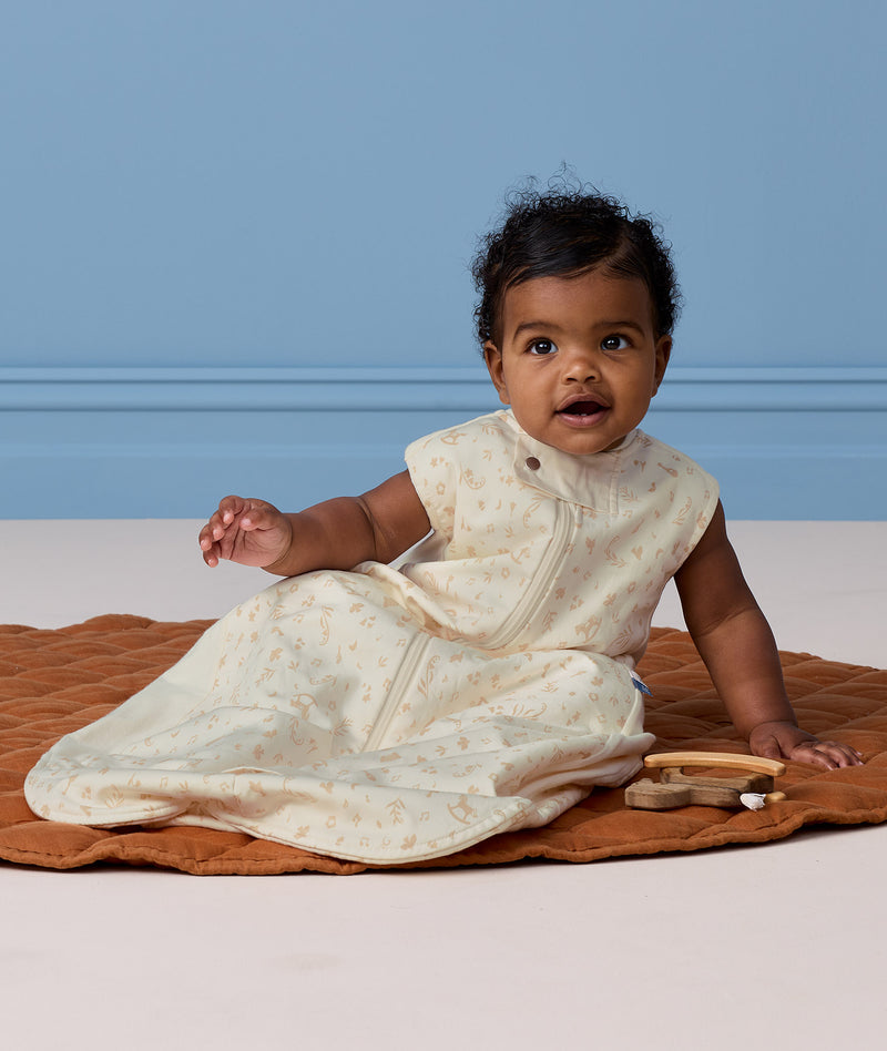 Baby sitting on a brown play mat wearing a cream patterned Sleeping Bag featuring a front zipper.