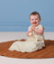 Smiling baby sitting on a brown play mat holding a rattle, wearing a light cream Premium Sleeping Bag 1.0 TOG.
