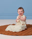 Smiling baby sitting on a brown play mat holding a rattle, wearing a light cream Premium Sleeping Bag 1.0 TOG.