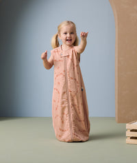 A young child stands smiling and gesturing with her hands while wearing a peach-colored sleeveless sleeping bag. The sleeping bag features a subtle all-over animal print in a slightly darker peach shade and has a front zipper for easy access.