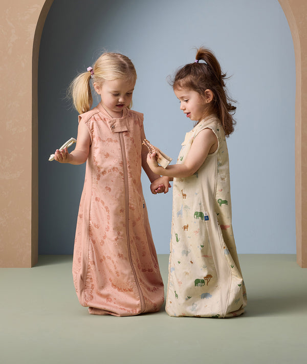 Two young children stand facing each other, holding hands and wooden toys while wearing sleeveless sleeping bags. The child on the left wears a peach-colored sleeping bag with a subtle animal print in a darker peach shade, and a front zipper. The child on the right wears a cream-colored sleeping bag with a playful, multicolored animal print. Both sleeping bags are loose-fitting and cozy, ideal for nighttime comfort.