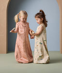 Two young children stand facing each other, holding hands and wooden toys while wearing sleeveless sleeping bags. The child on the left wears a peach-colored sleeping bag with a subtle animal print in a darker peach shade, and a front zipper. The child on the right wears a cream-colored sleeping bag with a playful, multicolored animal print. Both sleeping bags are loose-fitting and cozy, ideal for night time comfort.
