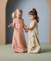 Two young children stand facing each other, holding hands and wooden toys while wearing sleeveless sleeping bags. The child on the left wears a peach-colored sleeping bag with a subtle animal print in a darker peach shade, and a front zipper. The child on the right wears a cream-colored sleeping bag with a playful, multicolored animal print. Both sleeping bags are loose-fitting and cozy, ideal for night time comfort.