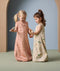 Two young children stand facing each other, holding hands and wooden toys while wearing sleeveless sleeping bags. The child on the left wears a peach-colored sleeping bag with a subtle animal print in a darker peach shade, and a front zipper. The child on the right wears a cream-colored sleeping bag with a playful, multicolored animal print. Both sleeping bags are loose-fitting and cozy, ideal for nighttime comfort.
