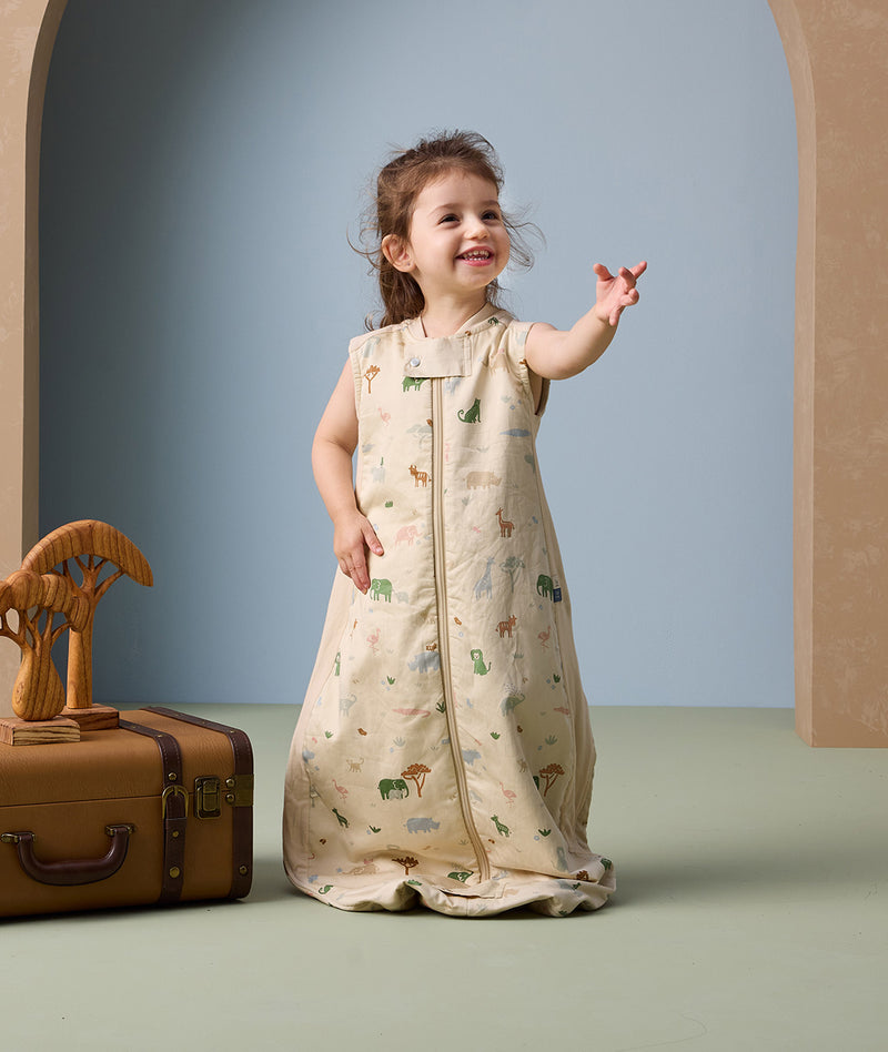 A young girl stands smiling and gesturing with her hand while wearing a cream-colored sleeveless sleeping bag. The sleeping bag features a colourful all-over animal print and has a front zipper for easy access.