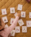 Toddler reaching for illustrated daily routine cards spread out on a mustard quilt, featuring activities like wash hands, tidy up, eat breakfast, play outside, brush teeth, put shoes on, and babysitter coming.