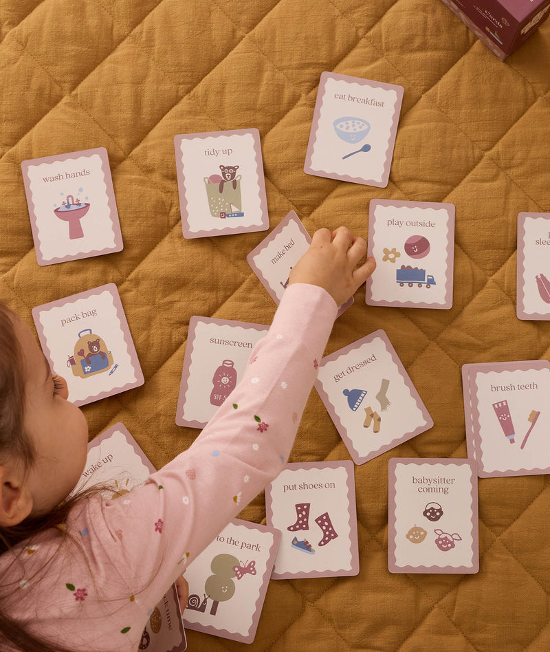 Toddler reaching for illustrated daily routine cards spread out on a mustard quilt, featuring activities like wash hands, tidy up, eat breakfast, play outside, brush teeth, put shoes on, and babysitter coming.