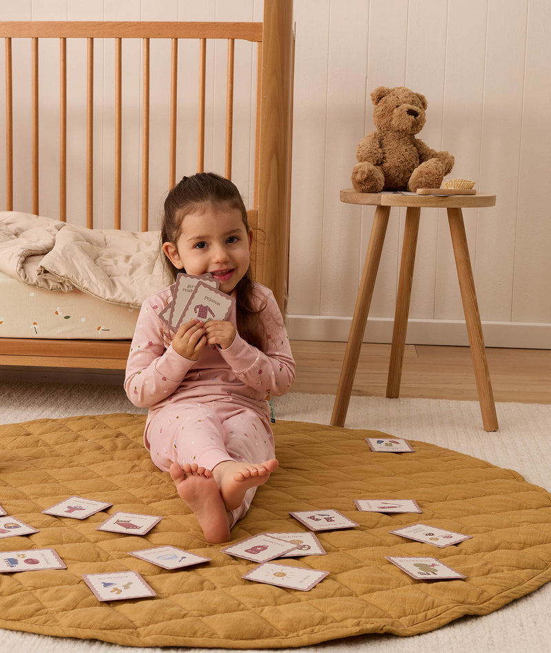 Toddler sitting on a mustard quilt in a nursery, holding illustrated daily routine cards, with more activity cards spread out around her on the floor near a wooden cot and a teddy bear on a side table.