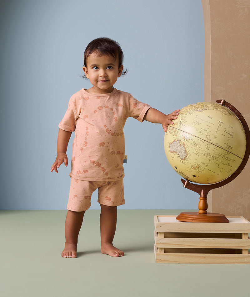 A toddler stands next to a vintage-style globe, gently touching it with one hand. The child is wearing a peach-colored short-sleeve pyjama set adorned with a subtle elephant print.