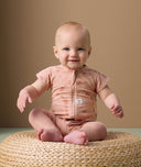 A smiling baby sits on a round woven cushion against a neutral beige background. The baby is wearing a short-sleeve layer in a soft peach color, featuring a subtle elephant print in a slightly darker peach tone.