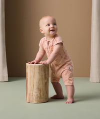 A baby stands while holding onto a wooden stump and is wearing a peach-colored short-sleeve layer with an all-over animal print. It features a zip-up front and is made from a soft, comfortable fabric, ideal for play and sleep.