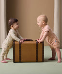 Two babies stand facing each other while holding onto a vintage-style brown suitcase. The baby on the left is wearing a cream-colored long-sleeve layer with a playful animal print. The baby on the right is dressed in a peach-colored short-sleeve layer featuring a subtle animal print in a darker peach tone.
