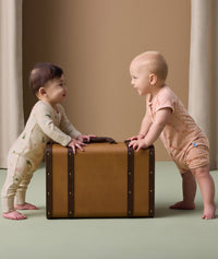 Two babies stand facing each other while holding onto a vintage-style brown suitcase. The baby on the left is wearing a cream-colored long-sleeve layer with a playful animal print. The baby on the right is dressed in a peach-colored short-sleeve layer featuring a subtle animal print in a darker peach tone.