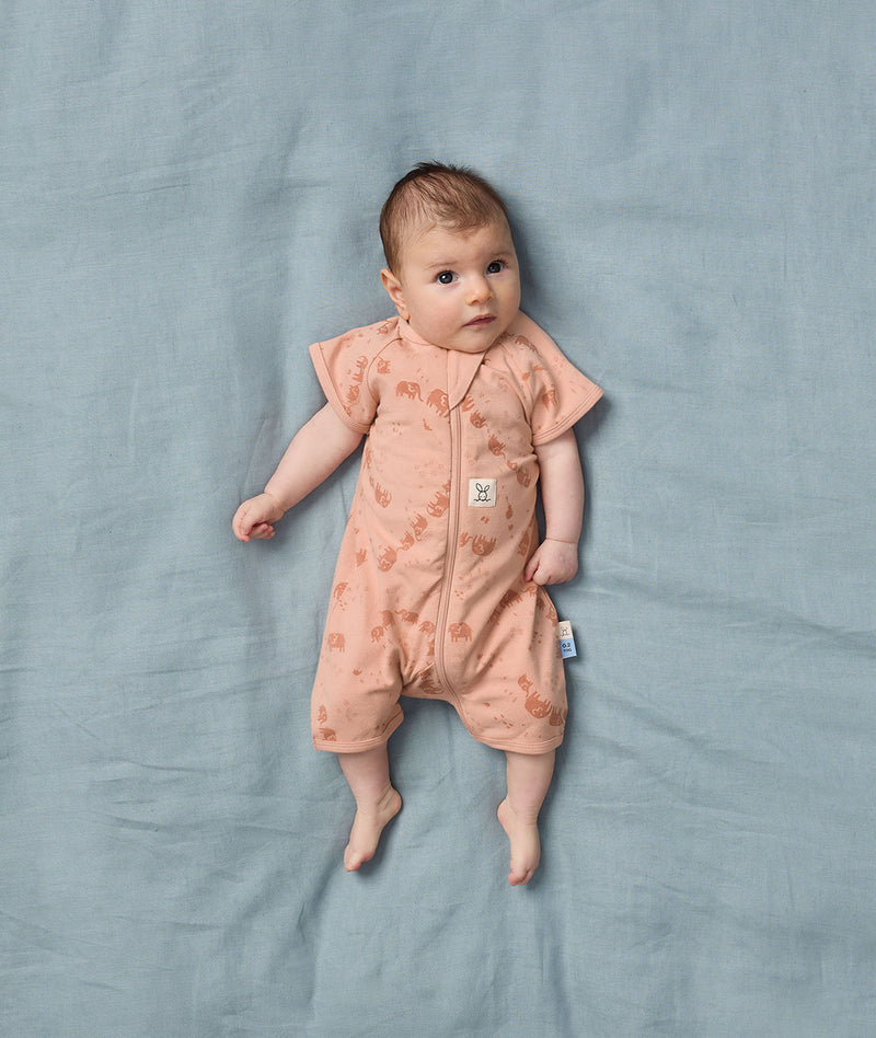 Baby is lying on a soft blue sheet, wearing an ergoPouch Short Sleeve Layer with elephant print, made from soft organic cotton gentle on baby's skin.