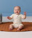Smiling baby sitting on a brown mat with hands raised, wearing a cream patterned Short Sleeve layer.