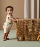 A baby stands while holding onto a large woven basket. The baby is wearing a cream-colored short-sleeve layer decorated with a playful safari animal print.