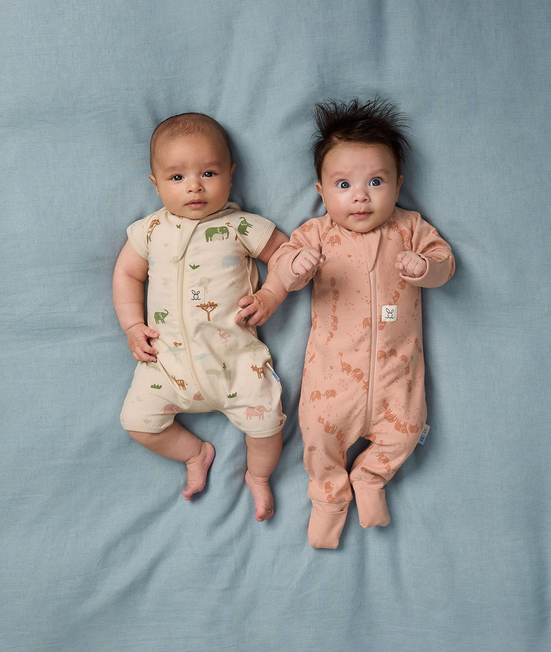 Two babies lying side by side wearing ergoPouch Short Sleeve Layer in Savannah and Long Sleeve Layer in Elephant Parade.