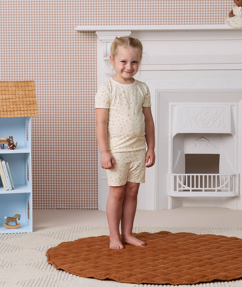 Young girl smiling and standing on a round brown rug, wearing a cream-colored Short Sleeve Pyjama set with a subtle pattern.