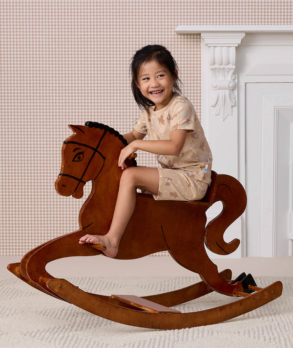 Preschooler wearing beige patterned Short Sleeved Pyjamas, riding a wooden rocking horse.