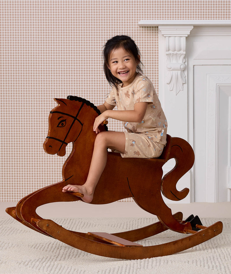 Preschooler wearing beige patterned Short Sleeved Pyjamas, riding a wooden rocking horse.