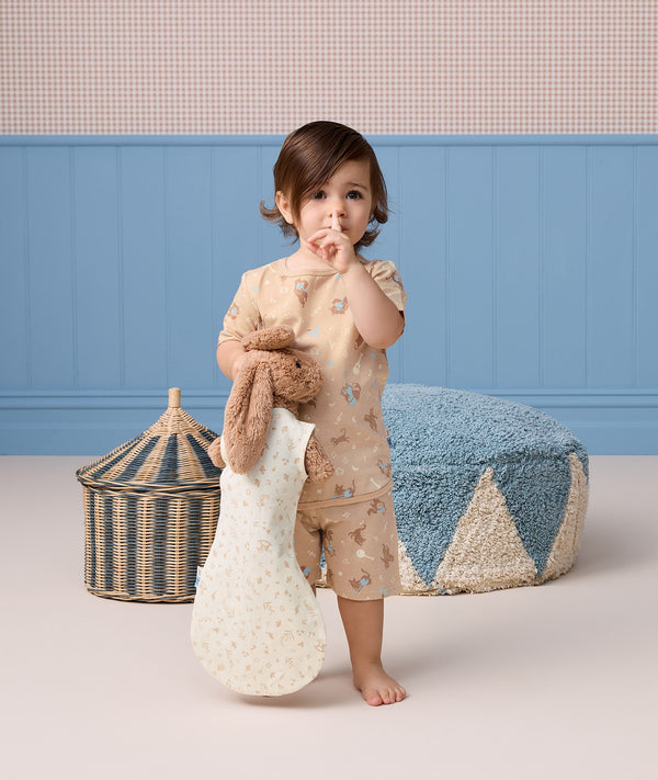 Toddler holding a stuffed bunny with a finger over their lips, wearing a beige patterned Short-Sleeve Pyjama set.