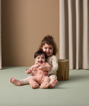 A young child sits on the floor holding a smiling baby in their lap. The older child is wearing cream-colored long-sleeve pyjamas with safari animal print in soft colors. The baby is dressed in a peach-colored long sleeve layer with elephant patterns.