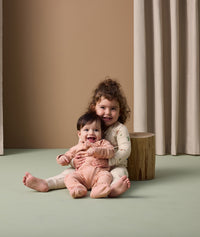 A young child sits on the floor holding a smiling baby in their lap. The older child is wearing cream-colored long-sleeve pyjamas with safari animal print in soft colors. The baby is dressed in a peach-colored long sleeve layer with elephant patterns.