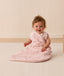 A smiling baby sitting on a white mat, wearing a pink ergoPouch Premium Sleeping Bag 1.0 TOG with a side zipper and a floral pattern.