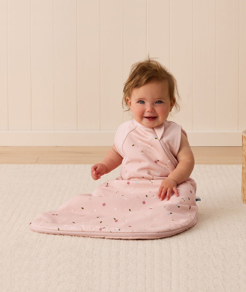 A smiling baby sitting on a white mat, wearing a pink ergoPouch Premium Sleeping Bag 1.0 TOG with a side zipper and a floral pattern.