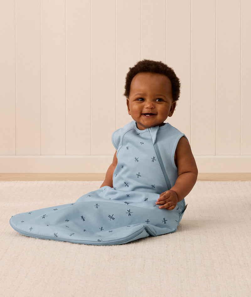 Smiling infant sitting on a white mat, wearing an ergoPouch Sleeping Bag in the Dragonflies print with a side zip.