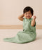 A happy baby sitting on a white mat, wearing an ergoPouch Side Zip Sleeping Bag 1.0 TOG Willow.