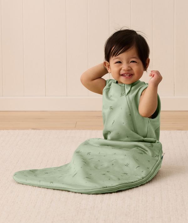 A happy baby sitting on a white mat, wearing an ergoPouch Side Zip Sleeping Bag 1.0 TOG Willow.