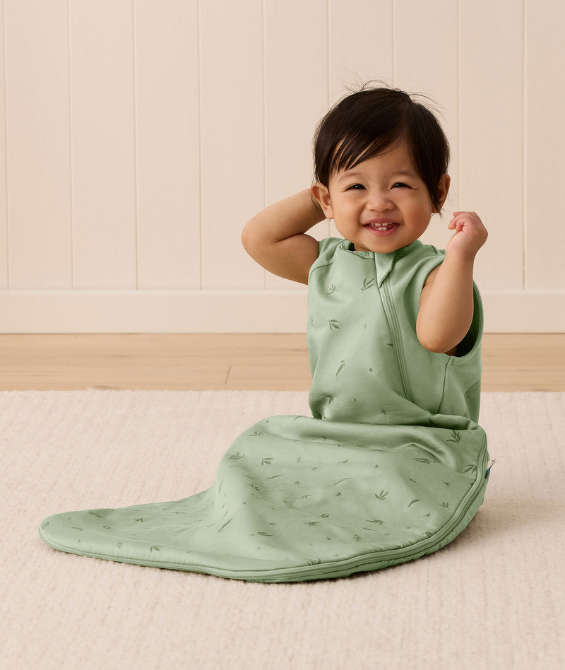 A happy baby sitting on a white mat, wearing an ergoPouch Side Zip Sleeping Bag 1.0 TOG Willow.