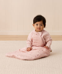 Baby sitting upright wearing a pink sleeping bag with small floral patterns, featuring arm openings and a side zipper.