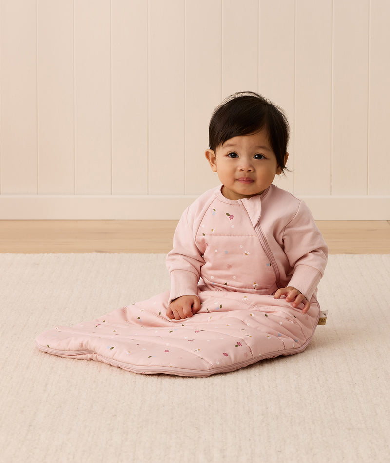 Baby sitting upright wearing a pink sleeping bag with small floral patterns, featuring arm openings and a side zipper.
