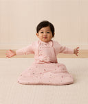 Baby sitting upright wearing a pink sleeping bag with small floral patterns, featuring arm openings and a side zipper.