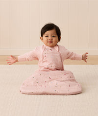 Baby sitting upright wearing a pink sleeping bag with small floral patterns, featuring arm openings and a side zipper.