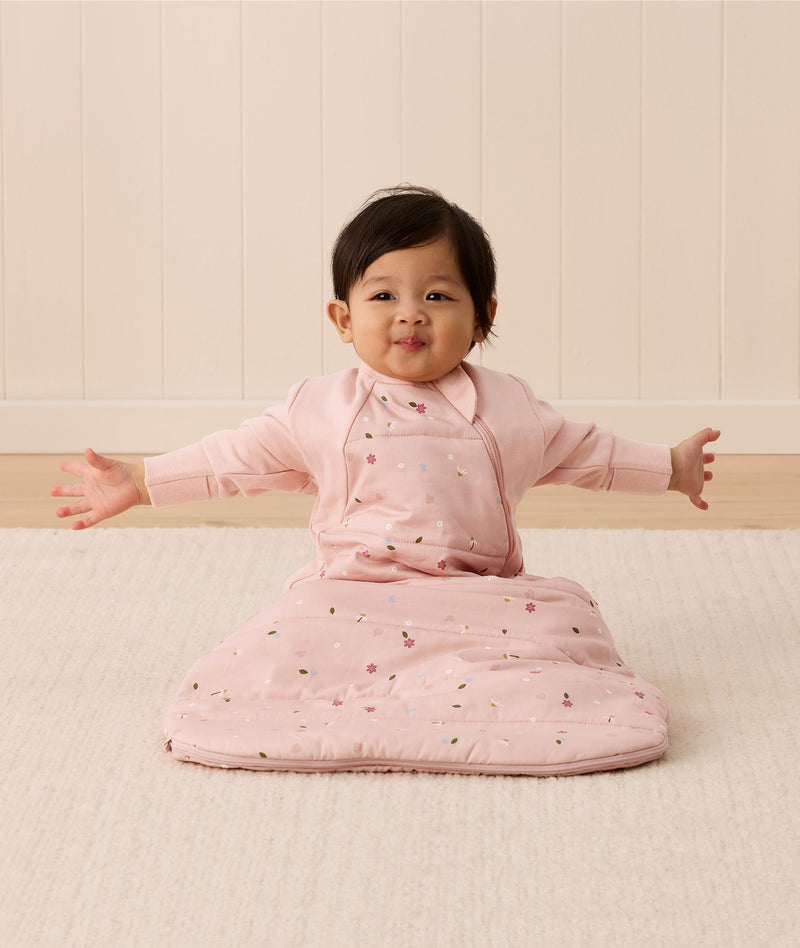 Baby sitting upright wearing a pink sleeping bag with small floral patterns, featuring arm openings and a side zipper.