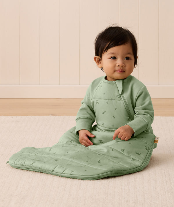 An infant sitting on a rug is wearing an ergoPouch Premium Sleeping Bag featuring side zippers.