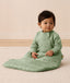 An infant sitting on a rug is wearing an ergoPouch Premium Sleeping Bag featuring side zippers.